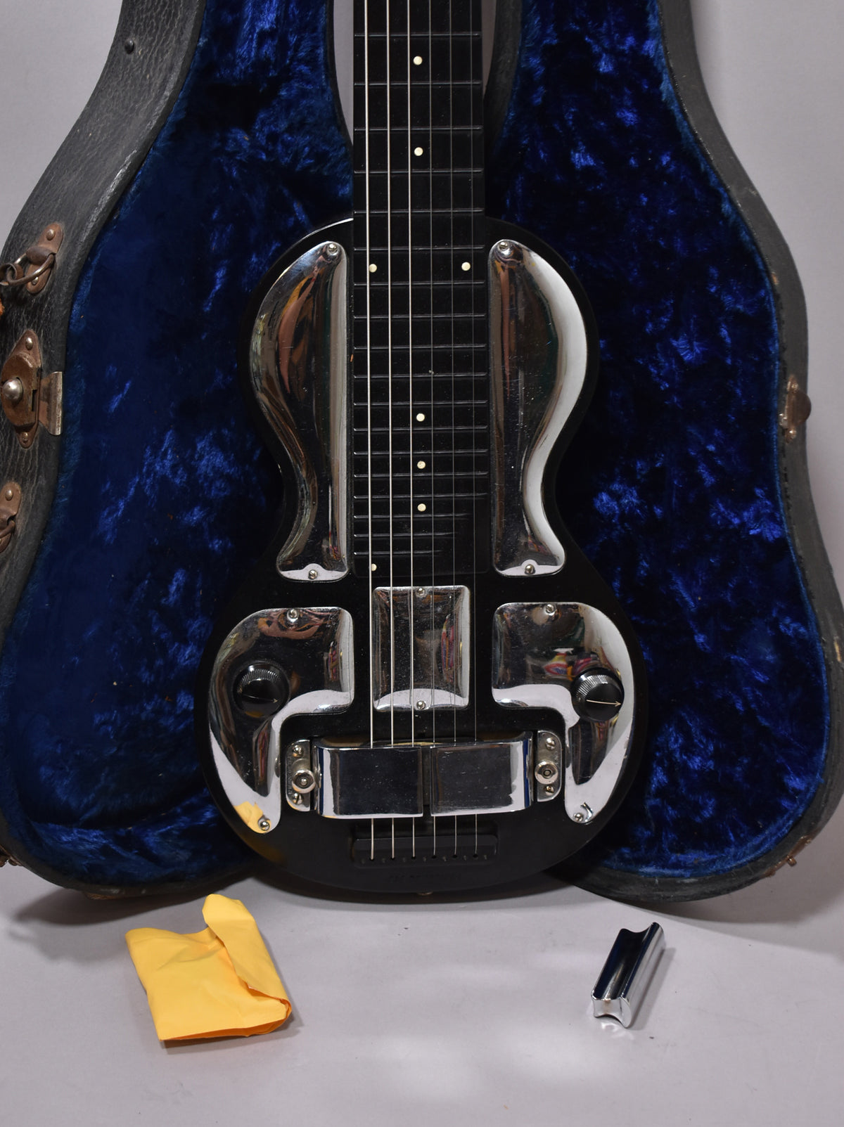 Black lap steel guitar with a chrome-plated body, single pickup, and a volume knob, resting in a blue velvet-lined case.