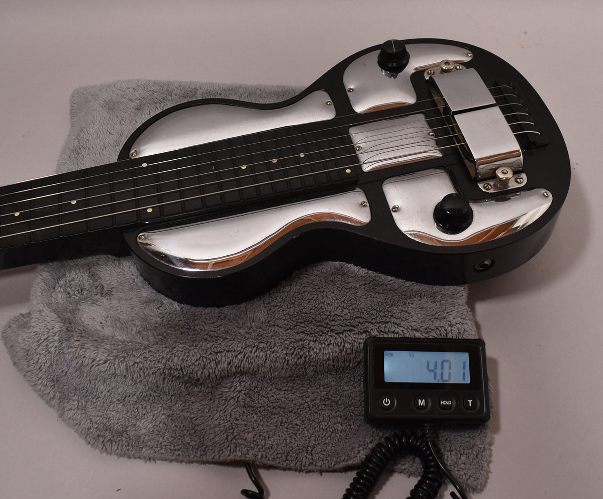 Black lap steel guitar with shiny chrome plates and hardware, showing some wear, resting on a gray fuzzy pad with a scale displaying "4.01".