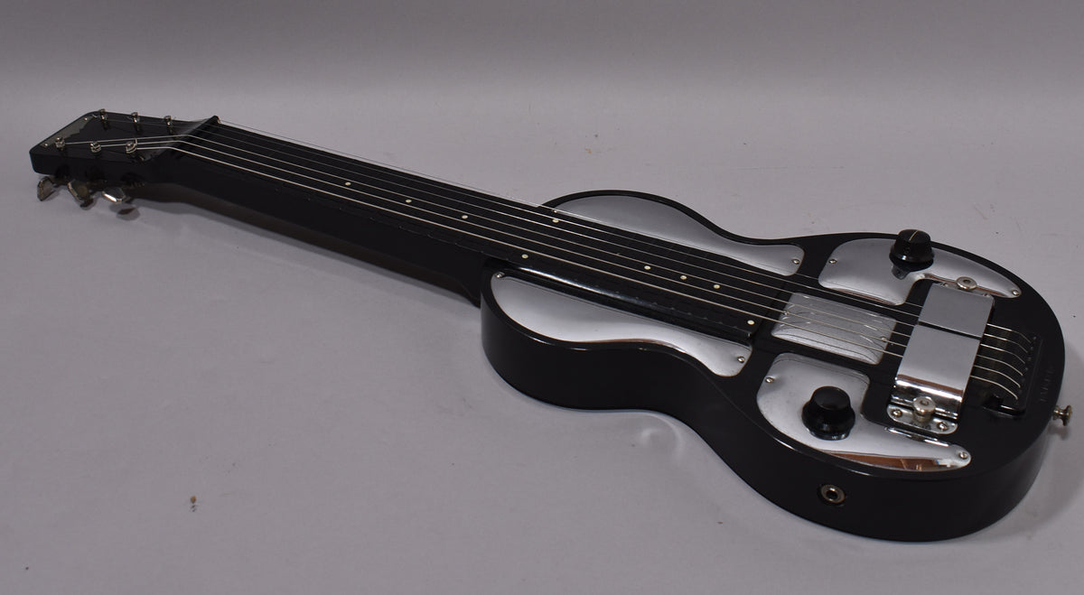 The black lap steel guitar is angled from the headstock, revealing its shiny chrome control plates, two black knobs, and output jack.