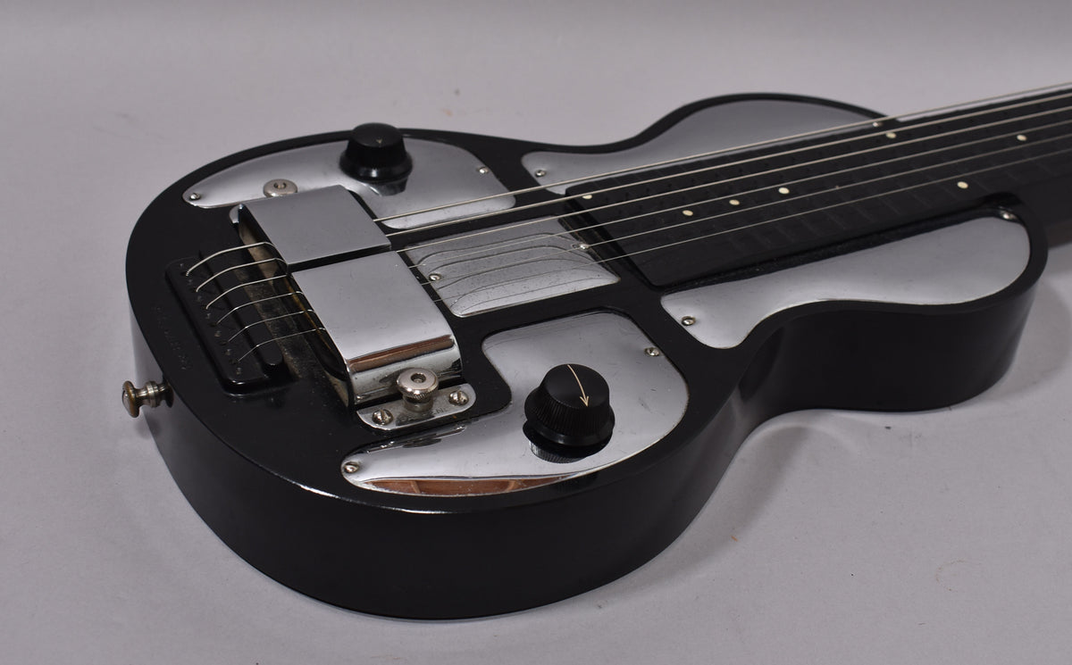 The black lap steel guitar is angled from the lower right, showing its shiny chrome control plates, two black knobs, and six strings.