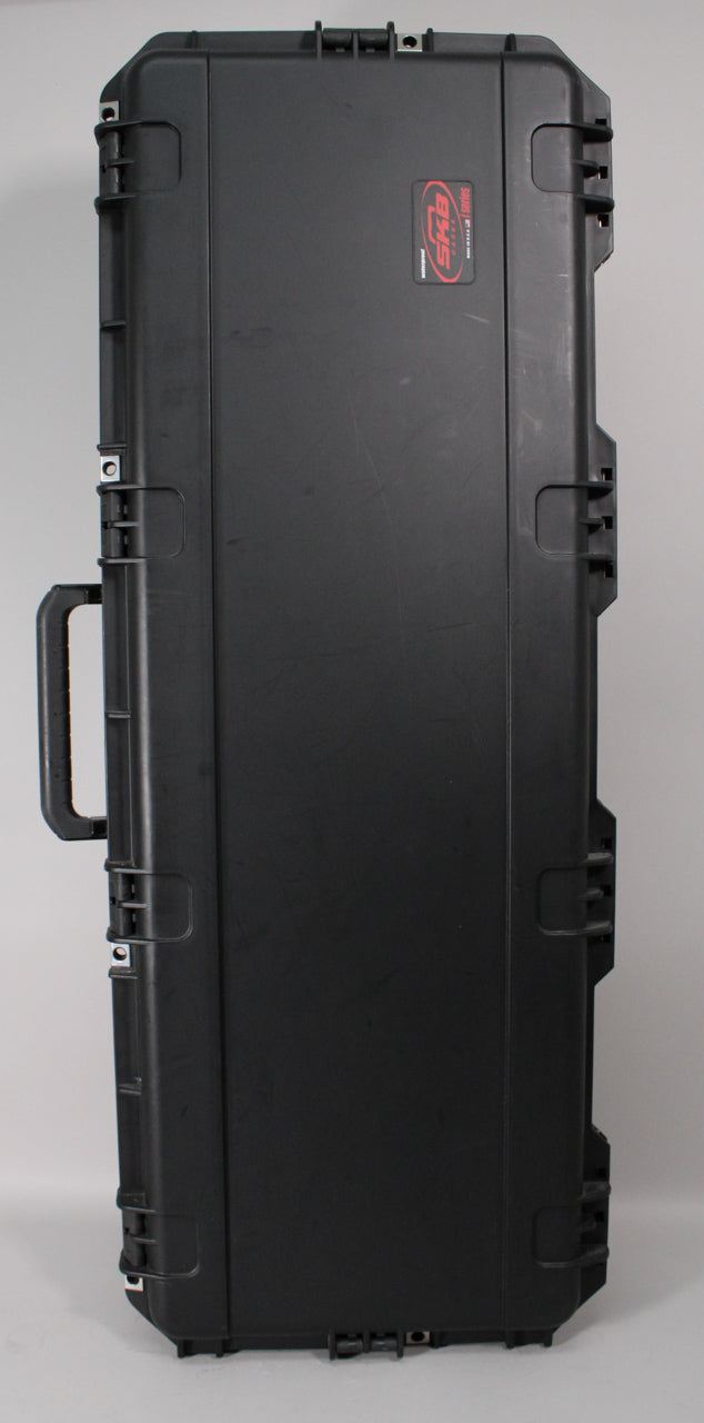A black hard case stands upright, showing its long side with a handle, six latches, and a red rectangular label.