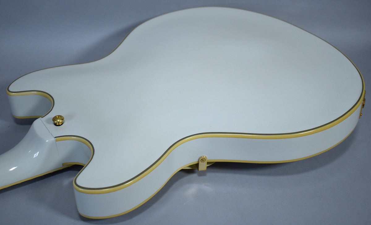 The back of a glossy white hollow-body electric guitar with gold binding, showing a gold strap button and light surface scuffs.