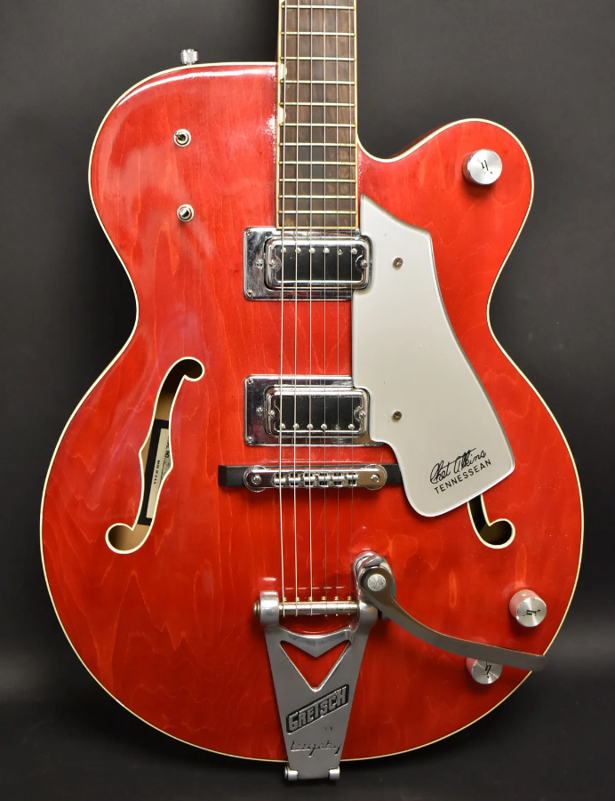 Full-body view of a cherry red hollow-body electric guitar with a Bigsby tremolo, two humbuckers, and a white pickguard.