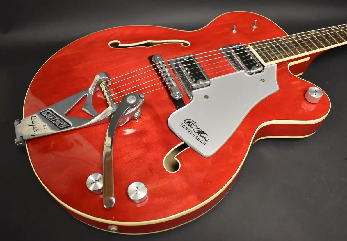 The cherry red hollow-body guitar, with a Bigsby vibrato and silver pickguard, is angled to show the body and part of the neck.