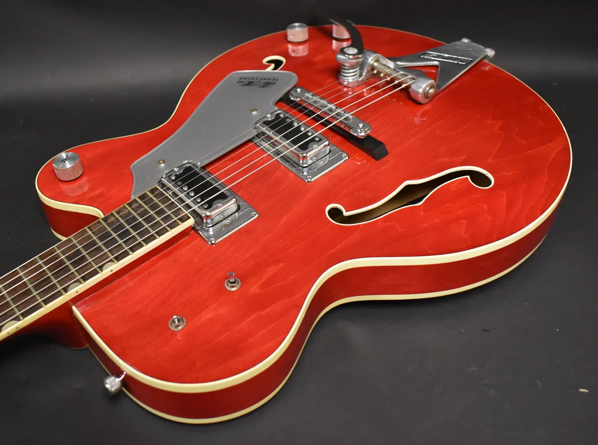 Cherry red hollow-body electric guitar, angled from the neck to the lower bout, showing two humbuckers, a Bigsby, and f-holes.