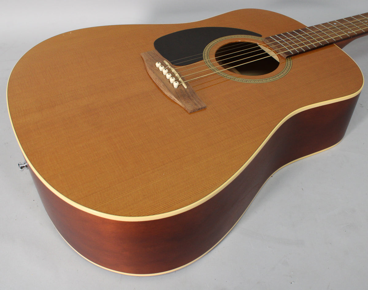 A left-handed acoustic guitar with a light cedar top, dark reddish-brown sides, and a black pickguard, viewed from the upper bout.
