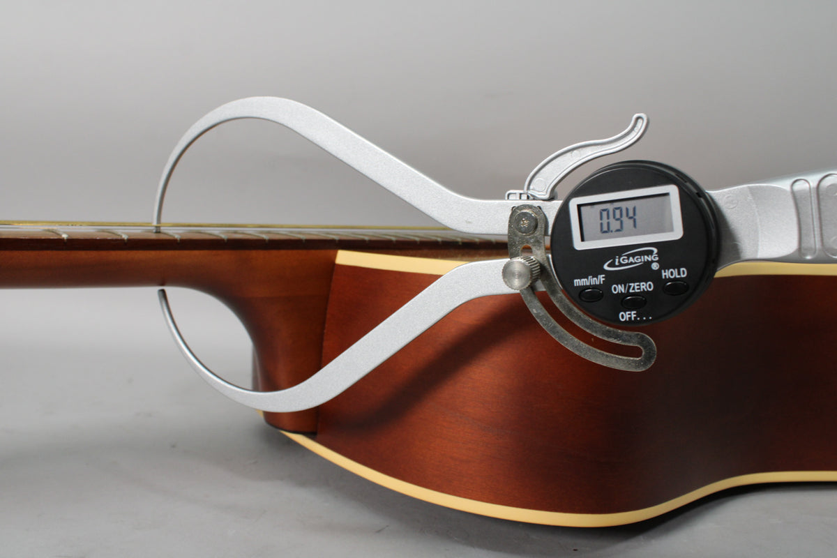 A digital caliper measures the string height at the 12th fret of the acoustic guitar's dark brown neck and body.