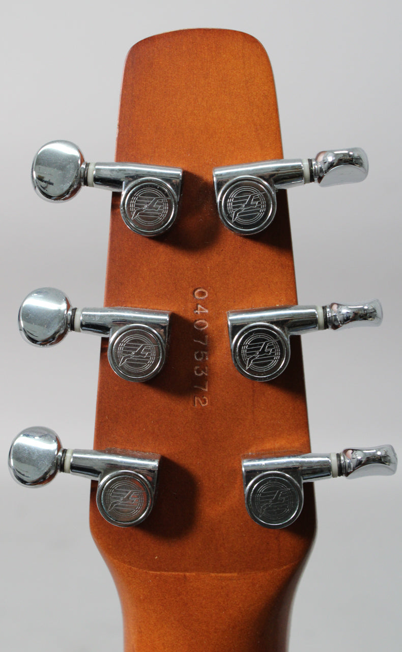 The back of a light brown headstock with six chrome tuners, three on each side, and a serial number stamped vertically.