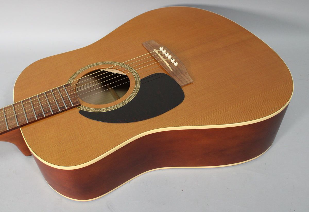 Left-handed acoustic guitar, angled view of the light brown top and dark brown sides, with a black pickguard and decorative soundhole rosette.