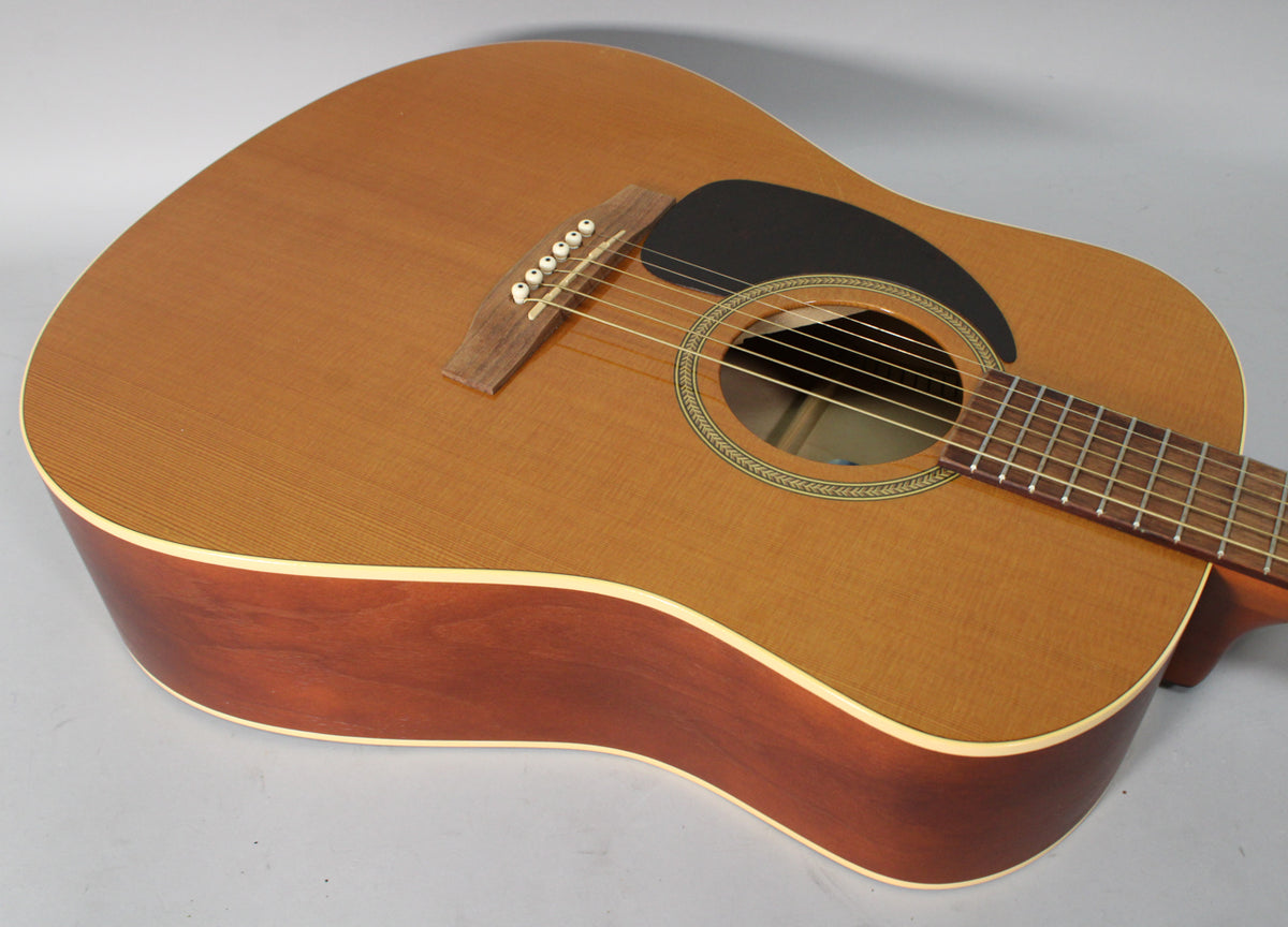 The left-handed acoustic guitar's light cedar top and dark mahogany sides are visible from above, with a black pickguard and ornate soundhole.