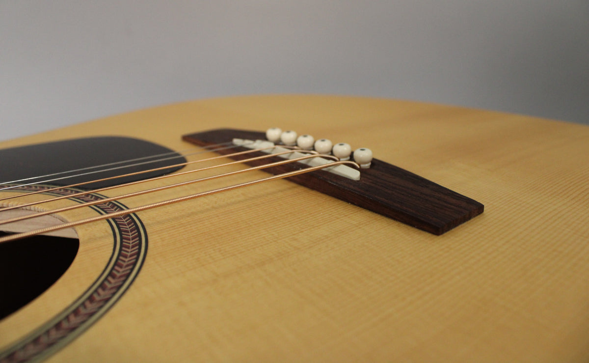 Close-up of the light-colored spruce top, showing the dark pickguard, rosette, bridge, and six white bridge pins with copper strings.