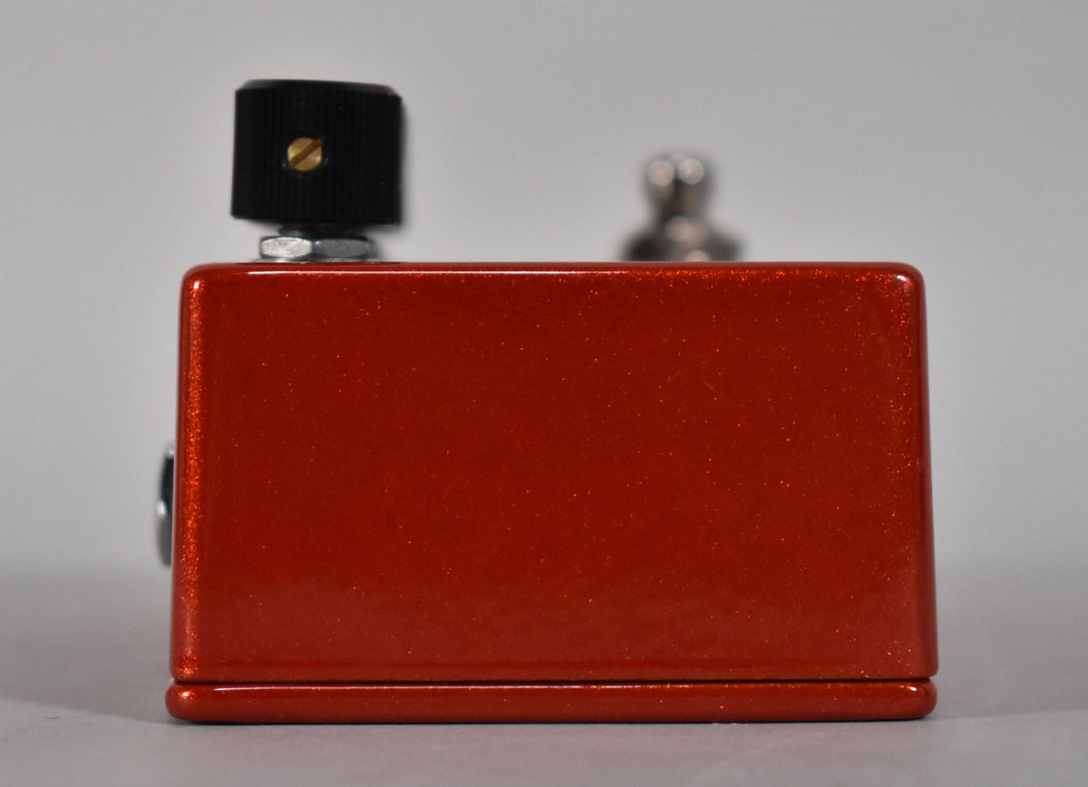 Side view of a metallic orange guitar pedal with a black knob on the left and a silver switch blurred in the background.