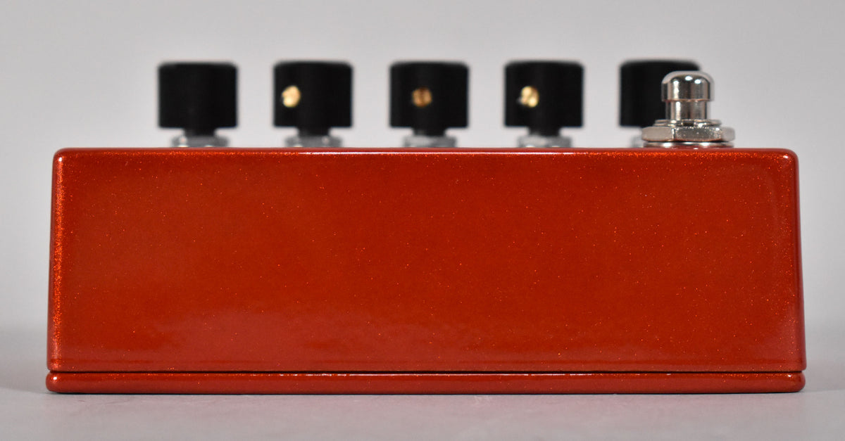The orange metallic sparkle pedal is viewed from the side, showing five black knobs with gold indicators and a chrome footswitch.