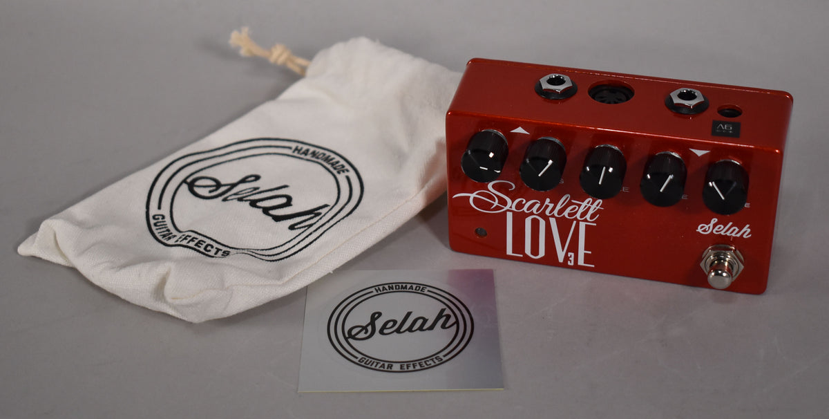 A glossy red guitar pedal with four black control knobs and a chrome footswitch, next to a white drawstring bag and sticker.