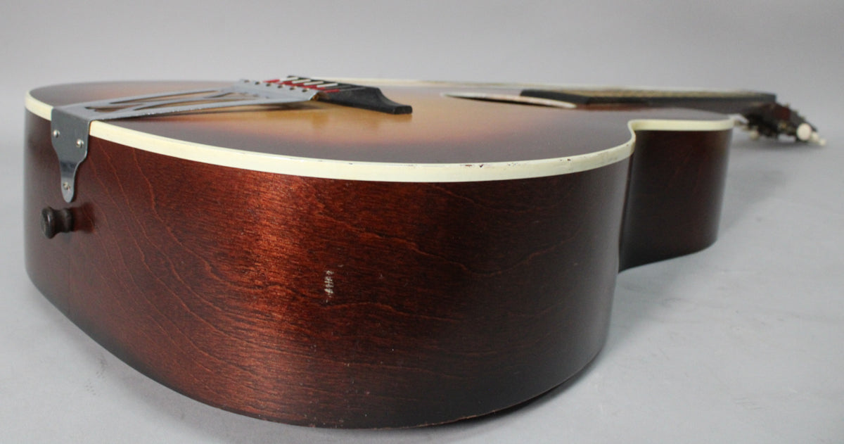The sunburst parlor guitar's lower bout and side, showing a strap button, tailpiece, and some scuffs on the dark brown finish.