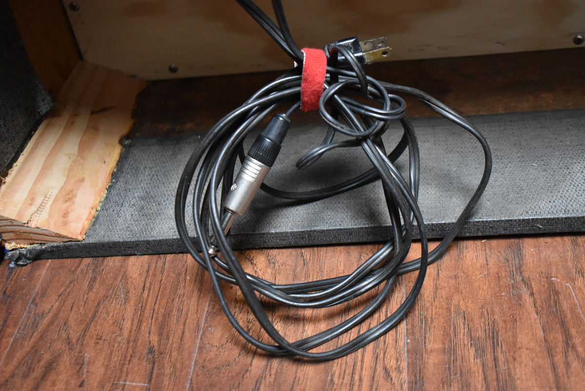 A coiled black cable with a 1/4-inch jack and a two-prong power plug, secured by a red velcro strap, rests on a wooden floor.
