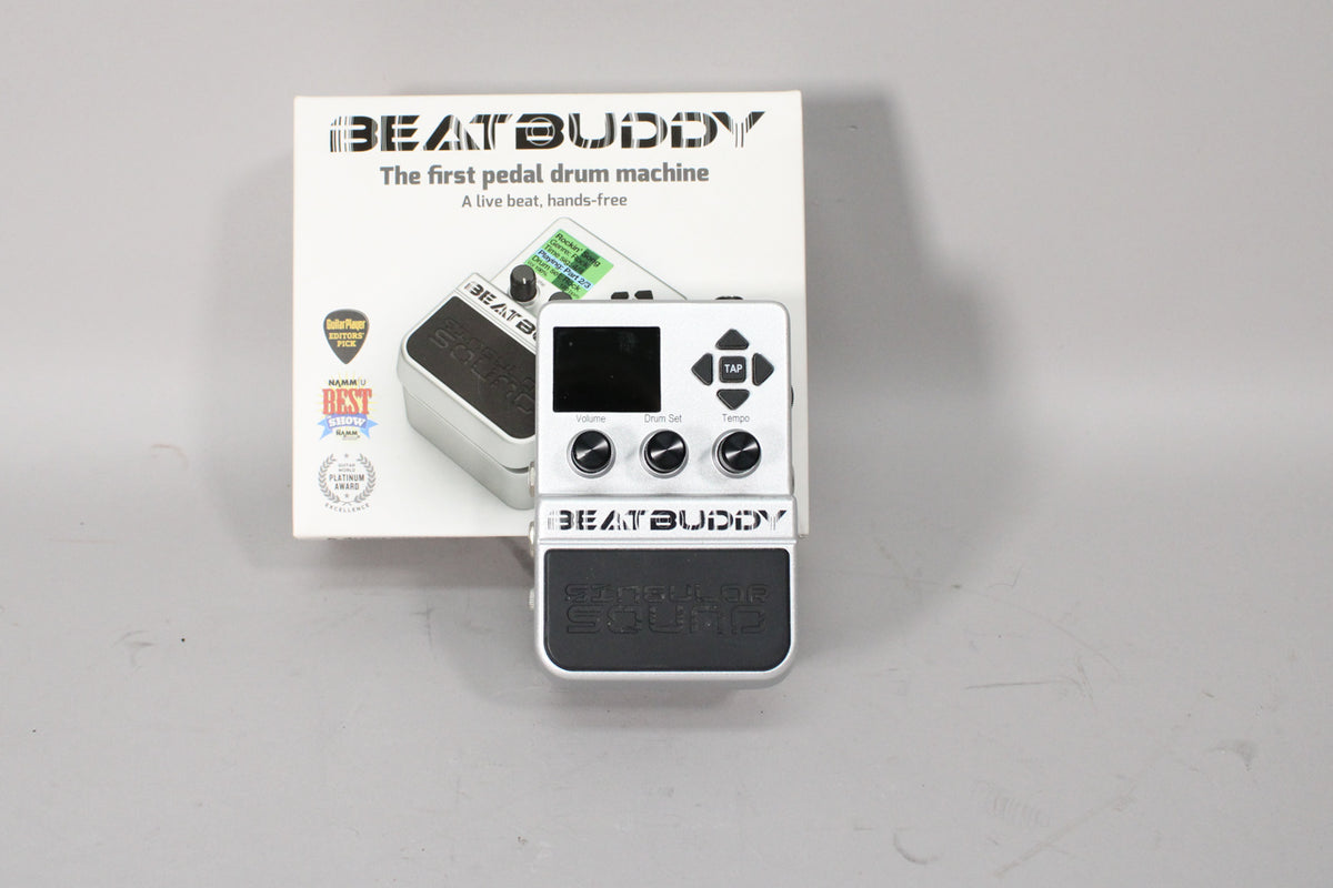 A silver-finished pedal with a black screen, three control knobs, a directional pad, and a large black footswitch, shown in its box.