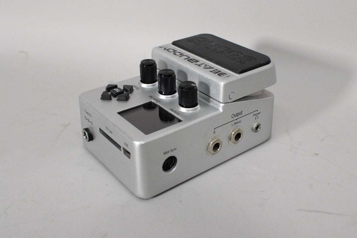A silver drum machine pedal, angled to show its top panel with three black knobs, a screen, and various ports on the sides.