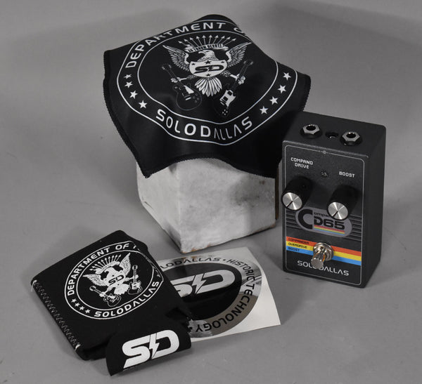 A dark gray pedal with two black knobs, a silver footswitch, and a colorful stripe, shown with branded accessories.