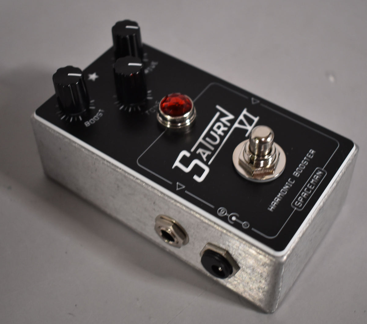 Angled view of a black pedal with a brushed aluminum base, showing three black knobs, a red jewel light, and a chrome footswitch.