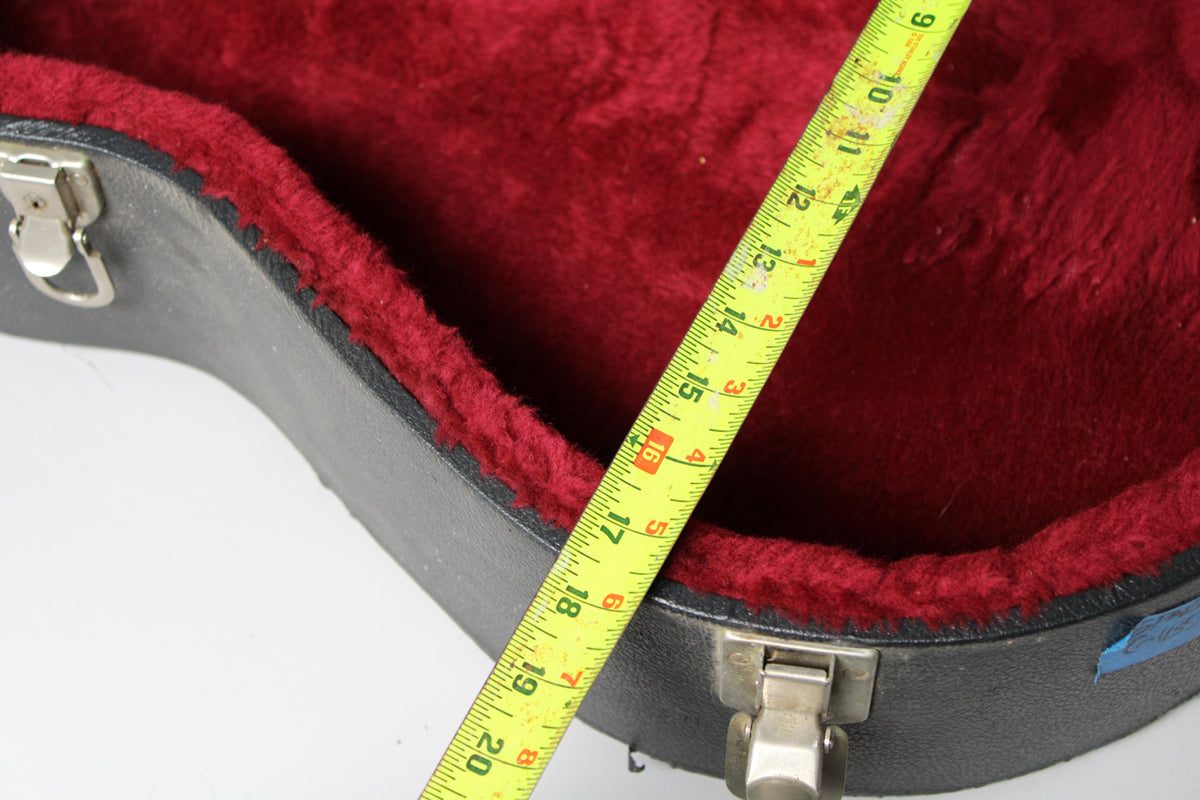 Open black archtop case with plush red lining, showing a tape measure across the opening, from 8 to 20 inches.