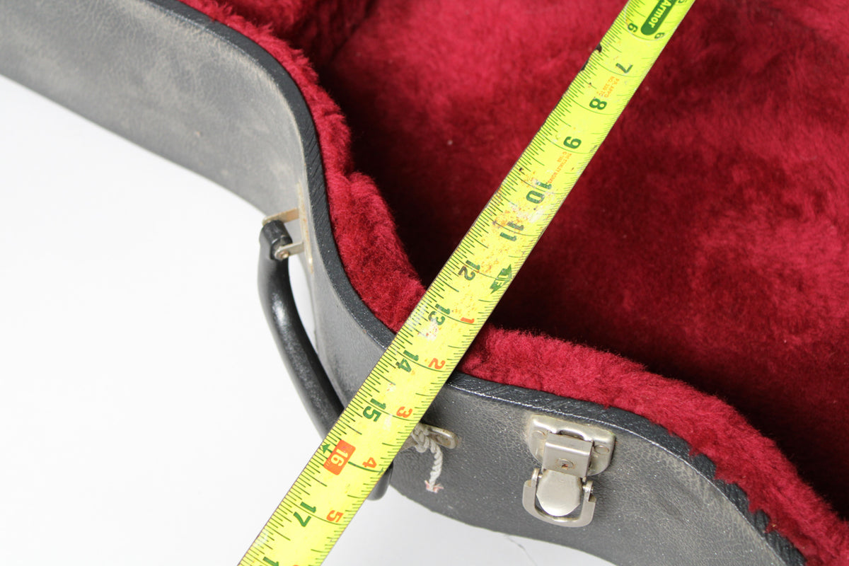 A black archtop case, open to show its red plush interior, with a yellow tape measure showing 17 inches.