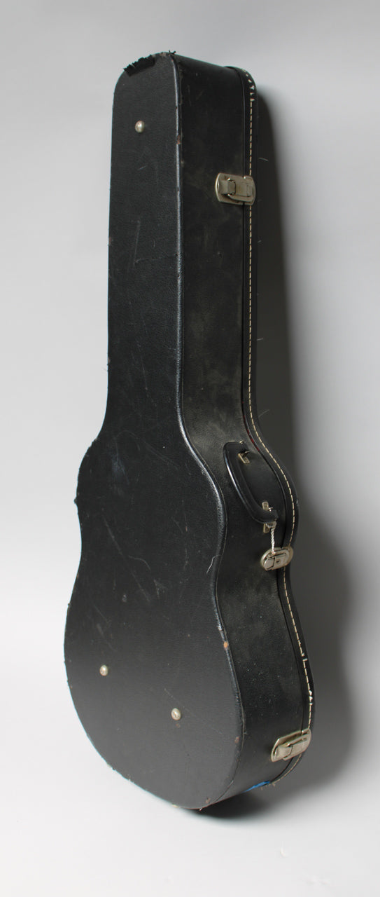 A black archtop guitar case, seen from the side, shows scuffs, worn edges, and three silver latches with white stitching.