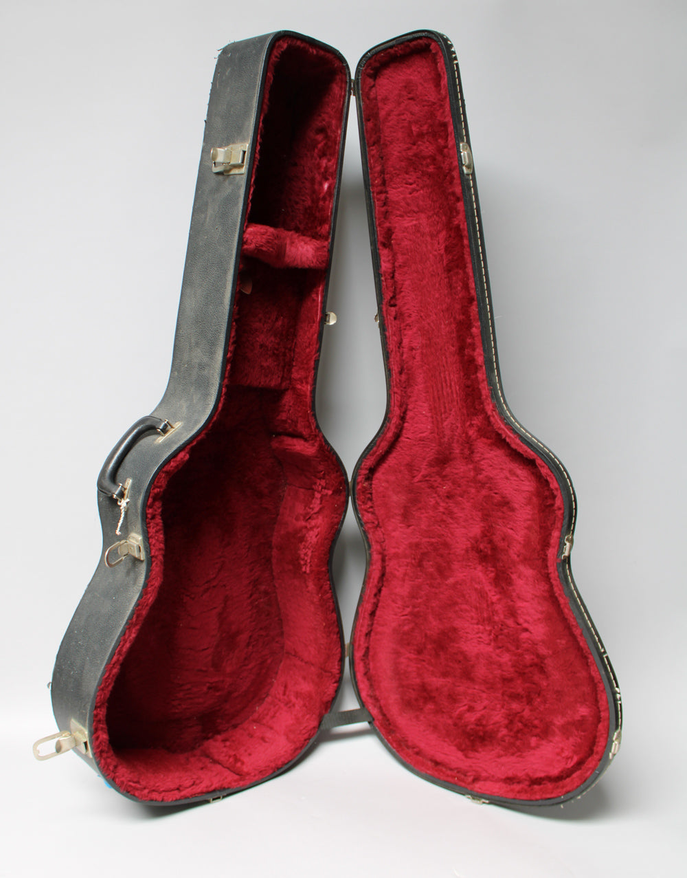 Open black archtop case with worn red plush lining, showing a small accessory compartment and tarnished brass latches.