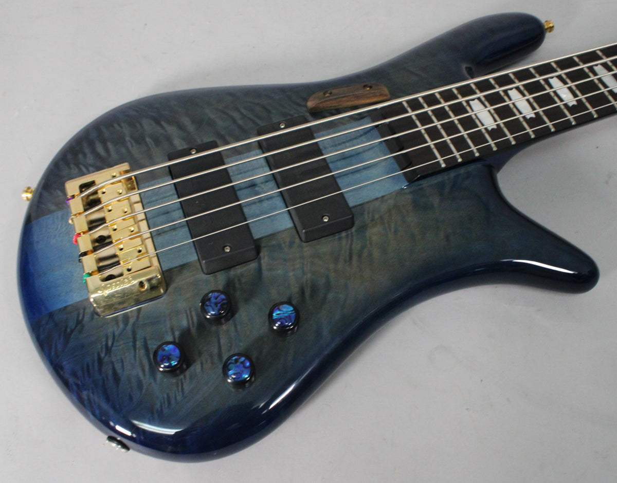Close-up of a blue fade gloss bass guitar, angled from the lower left, showing the gold bridge, black pickups, and four iridescent blue control knobs.