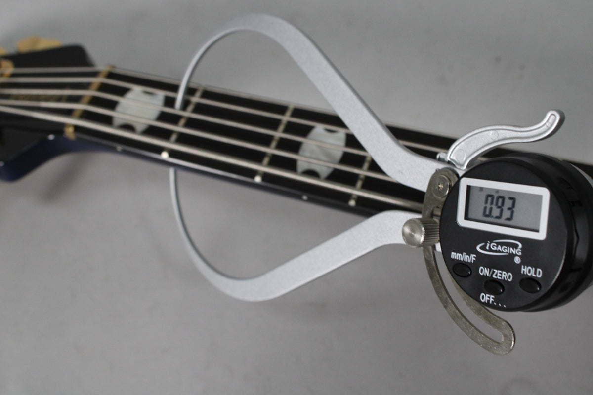 A digital caliper measures the bass's dark blue neck and fretboard with white block inlays, displaying "0.93" on its screen.