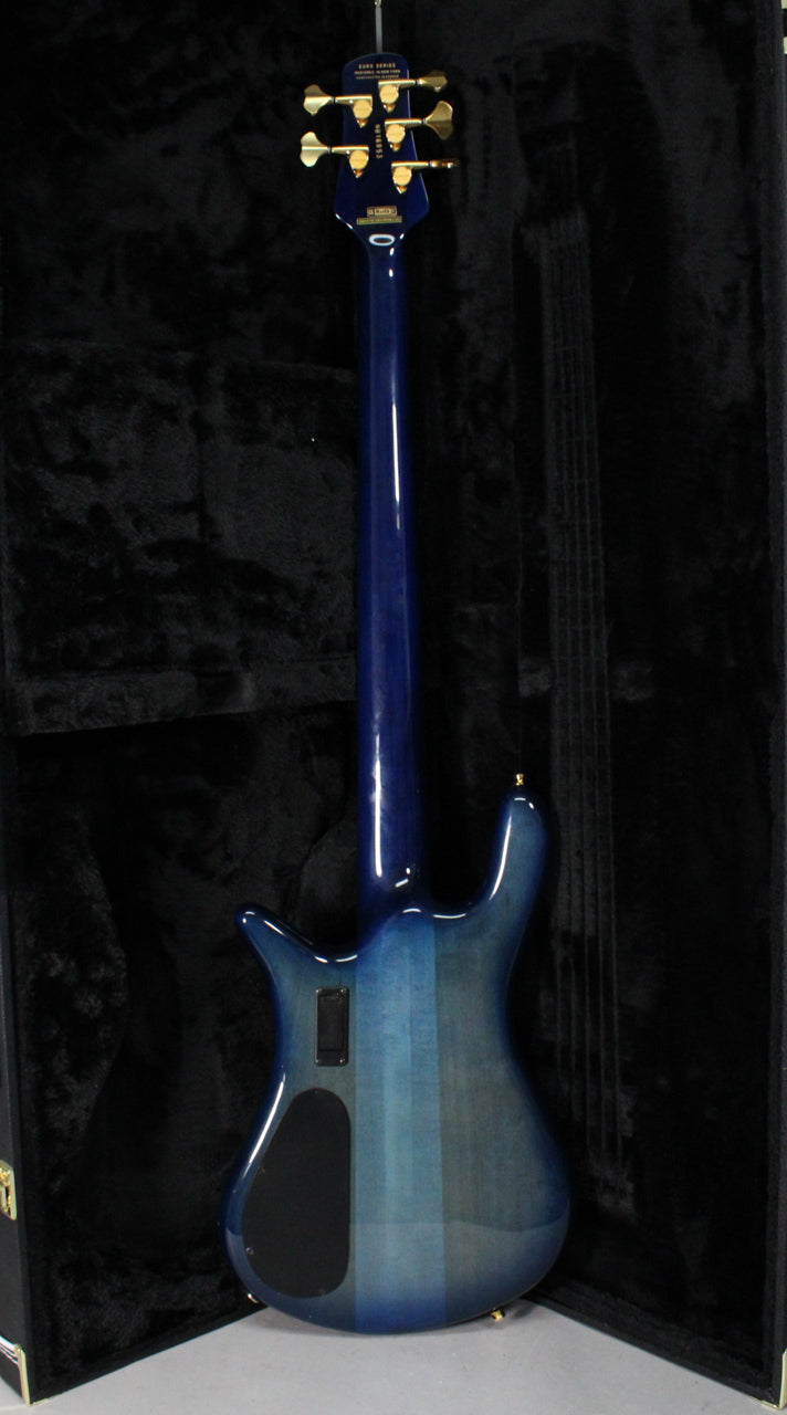 The bass guitar's back is shown, revealing a glossy blue fade finish, gold tuners, and a black control cavity cover.