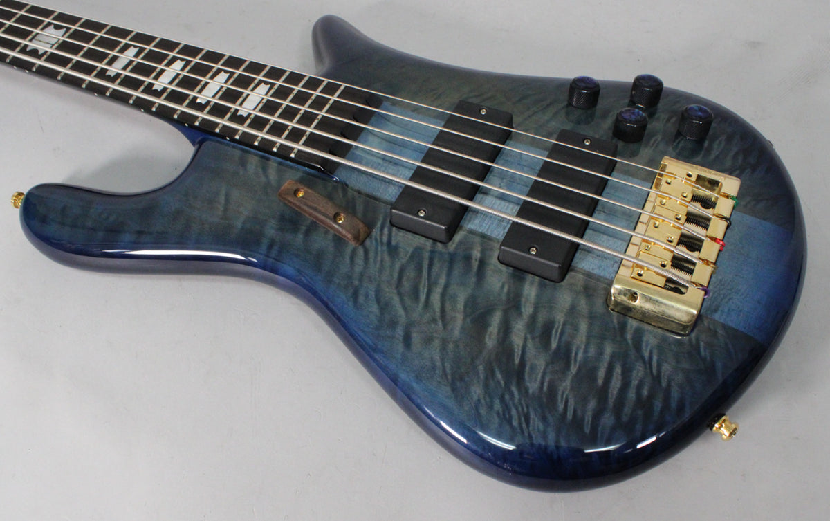 The bass guitar's blue fade gloss body has a wavy wood grain, with two black pickups, a gold bridge, and four dark control knobs.