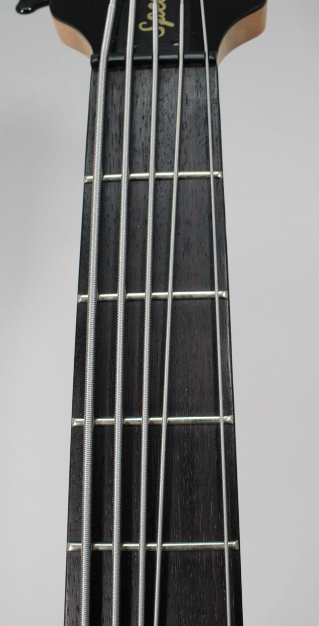 Close-up of the bass's dark fretboard and five silver strings, showing the first few frets and a light wood neck joint.