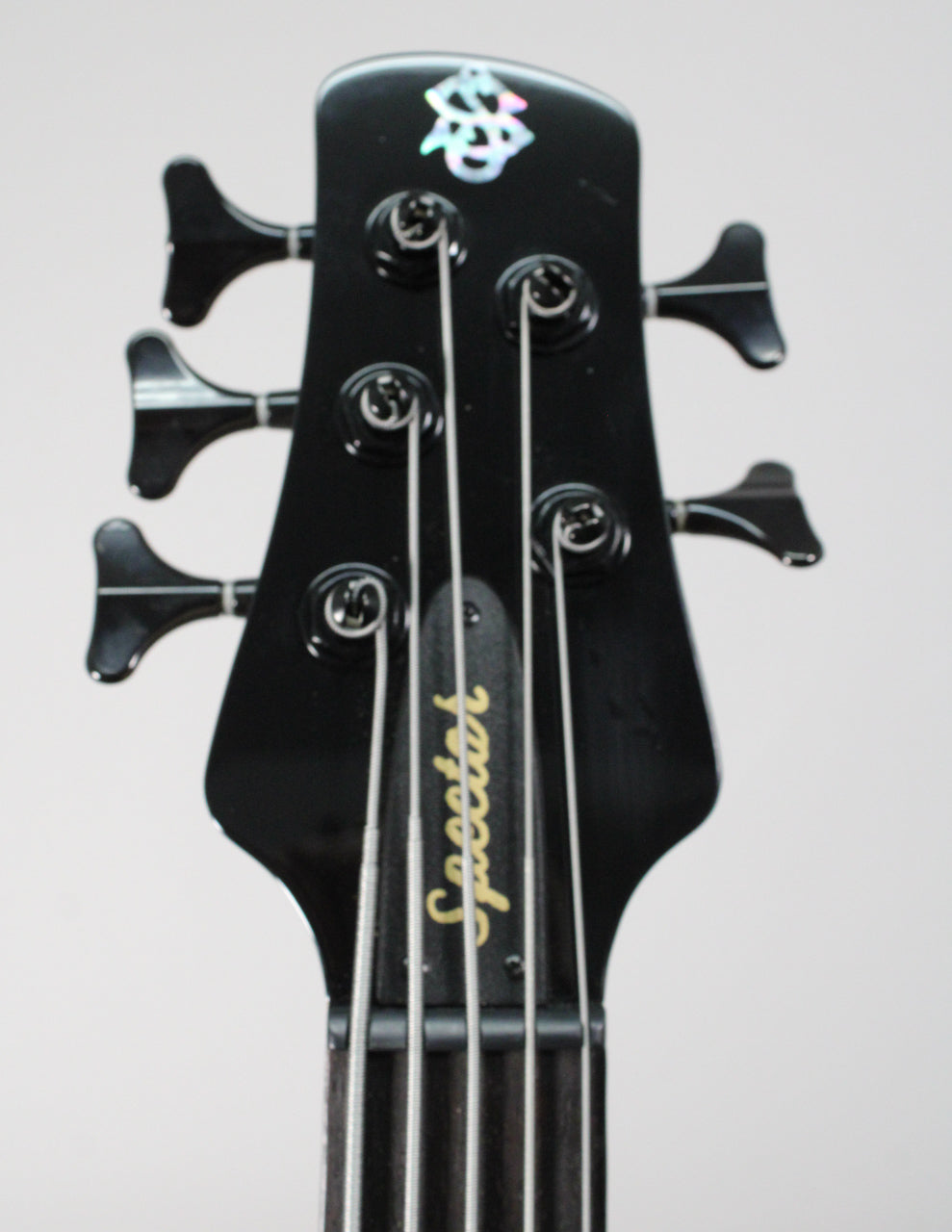The black gloss headstock of the 5-string bass, with black tuners and a pearlescent logo, is centered from above.
