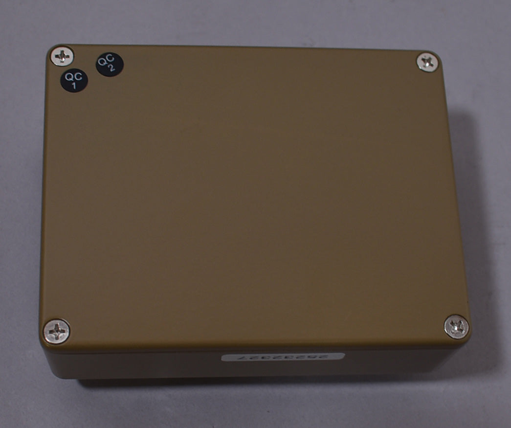 The pedal's tan enclosure is viewed from above, showing four silver Phillips head screws and two small black QC stickers.