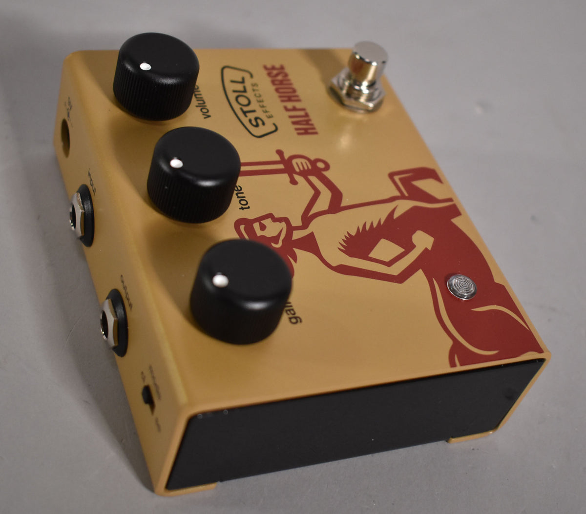 An angled view of the tan pedal with a red warrior graphic, three black knobs, a chrome footswitch, and two input jacks.