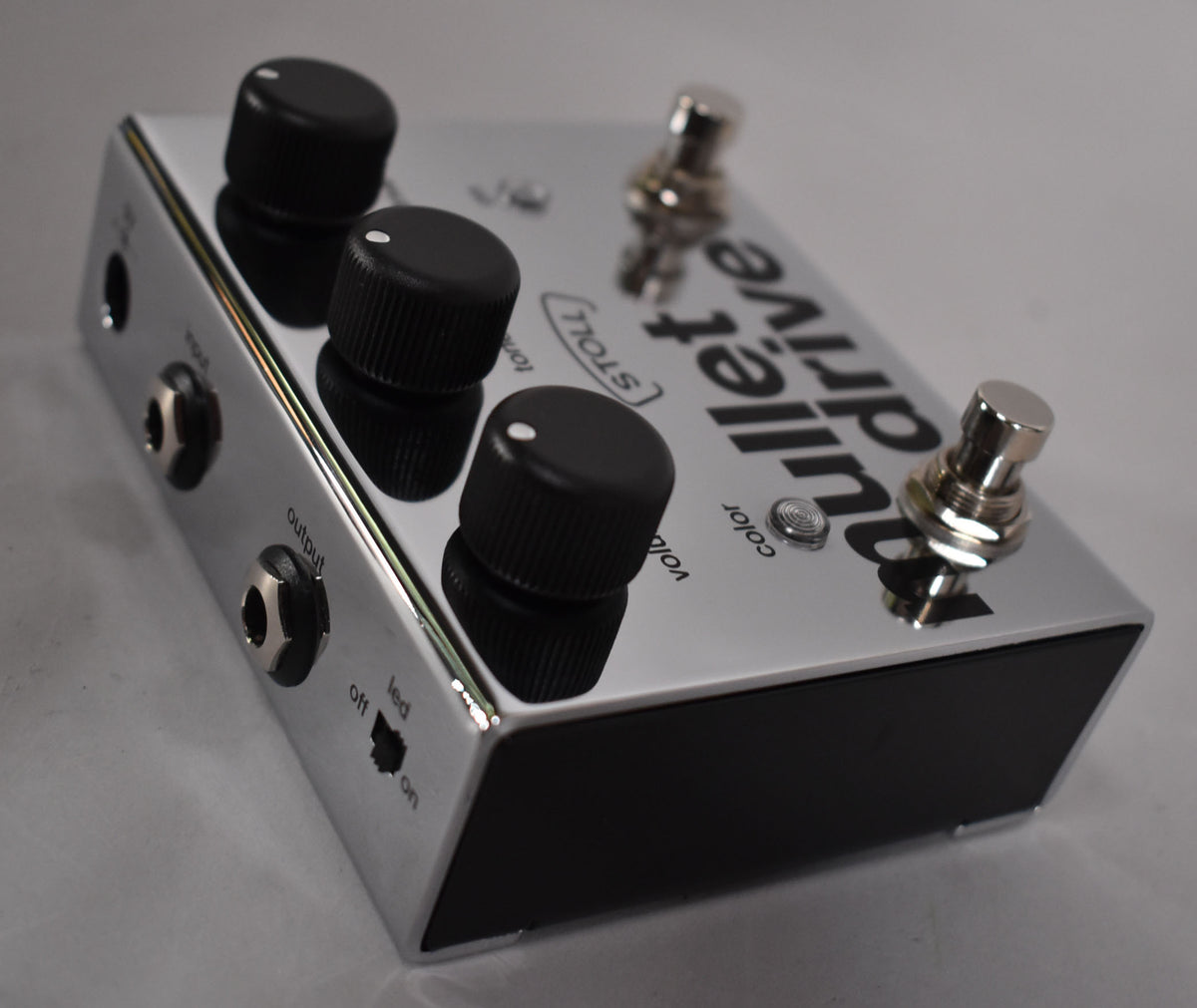 A chrome guitar pedal with three black control knobs, two footswitches, and input/output jacks, viewed from the upper left.