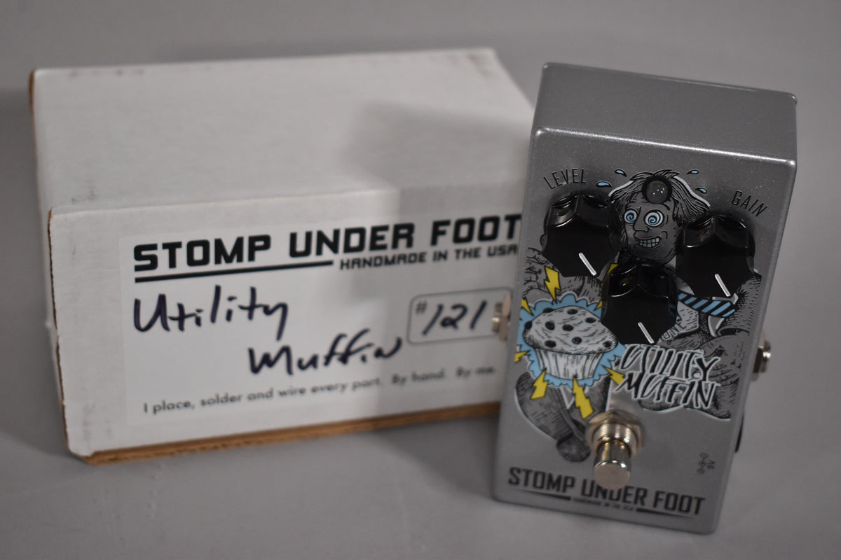 A silver fuzz pedal with black knobs and a cartoon graphic of a crazed scientist and a muffin, next to its white box.
