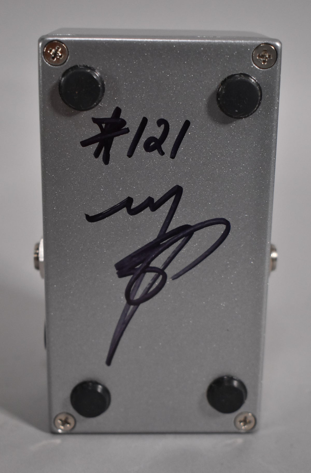 The bottom of a silver sparkle pedal with four black rubber feet, four silver screws, and black marker writing.