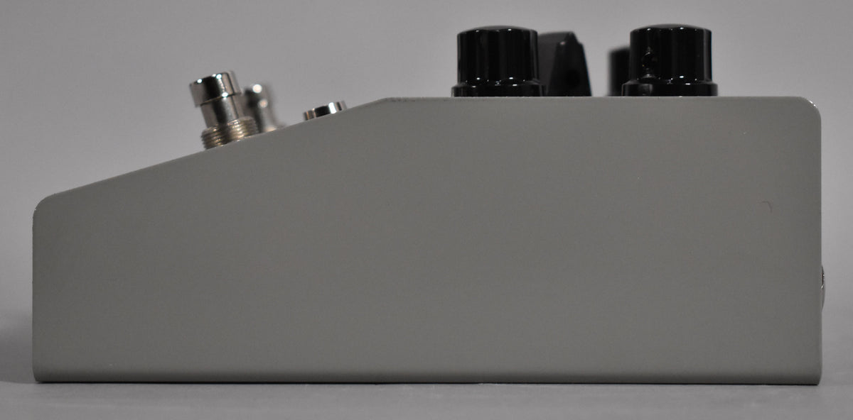 The gray pedal is viewed from the right side, showing two chrome footswitches and two black control knobs on the angled top surface.