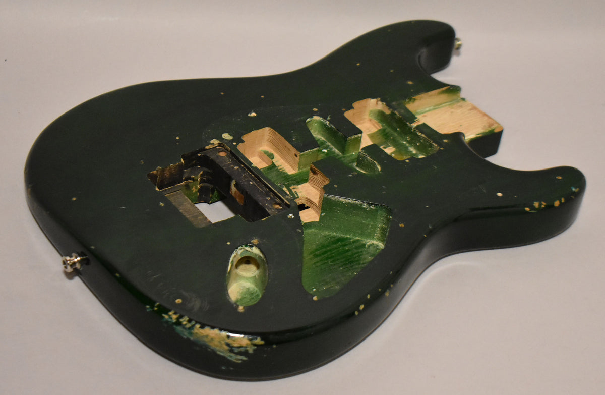 Dark green S-style guitar body, angled from above, showing the HSH routing, Floyd Rose cavity, and strap buttons, with paint chips.