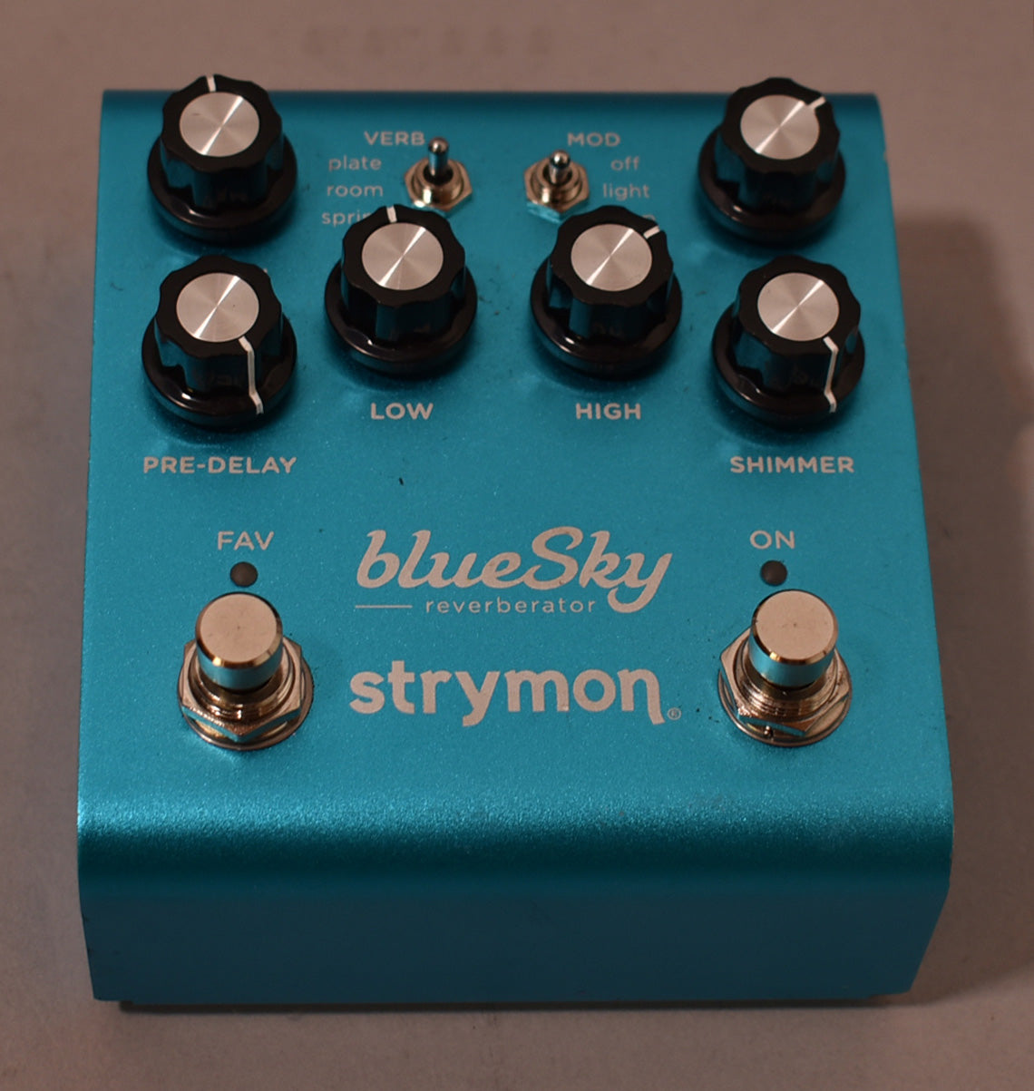 A top-down view of a metallic teal guitar pedal with six black knobs, two toggle switches, and two silver footswitches.