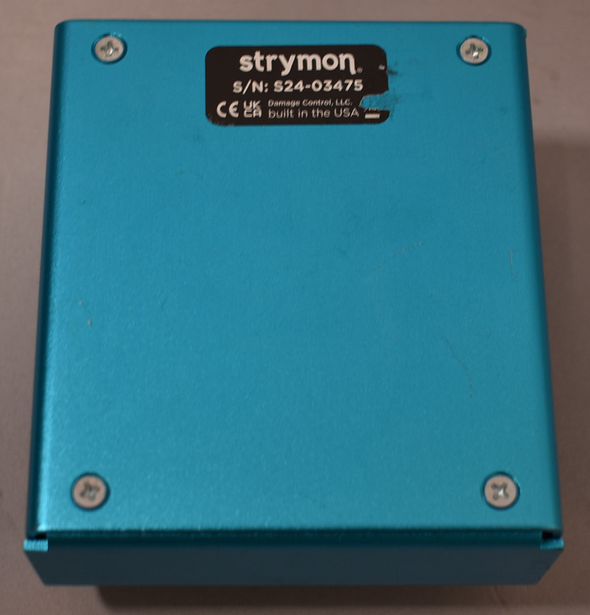 The pedal's teal underside is visible, secured by four silver Phillips head screws, with a black serial number sticker.