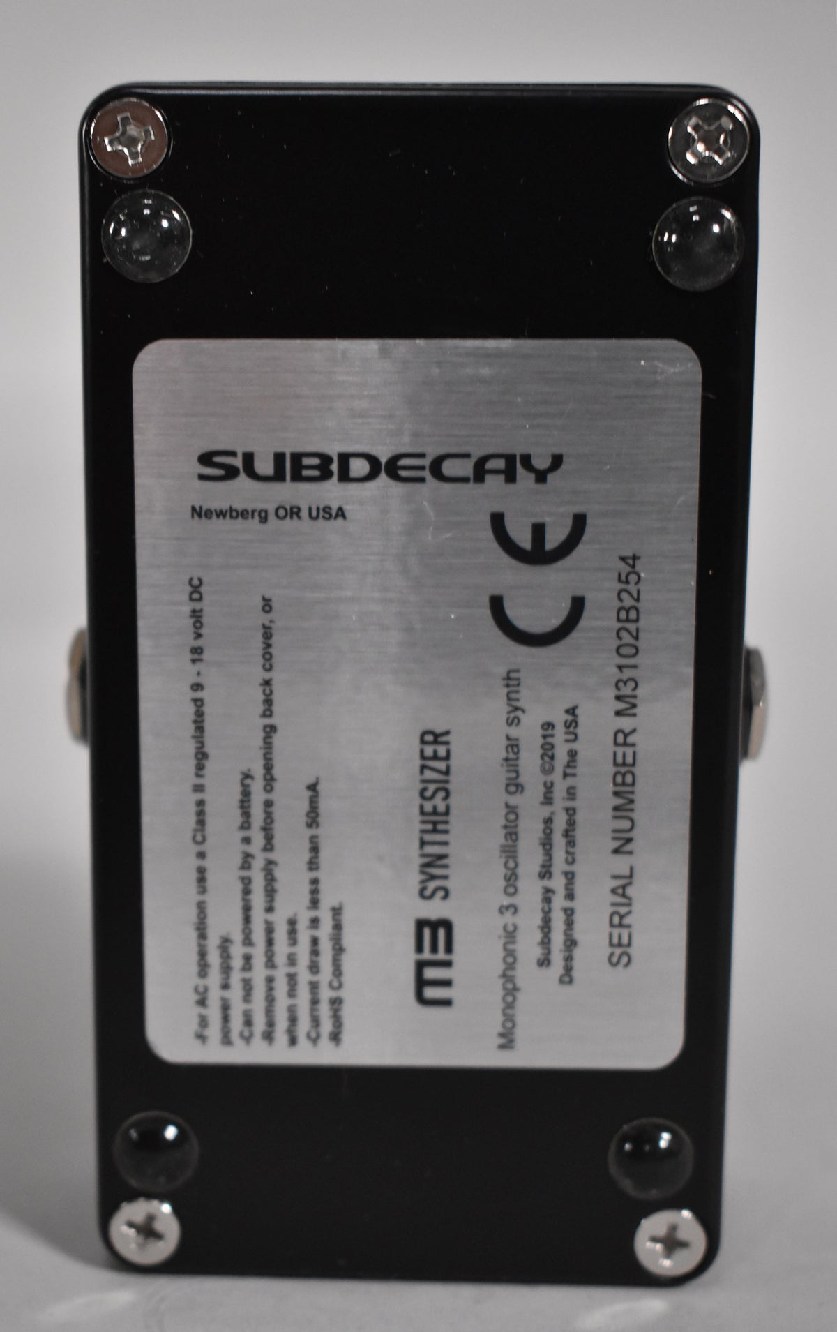 The black pedal is viewed from the back, showing a silver label with text and four visible screws and rubber feet.