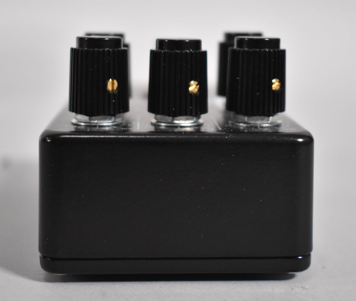 Side view of the black synth pedal, showing its textured finish, four black ridged knobs with gold indicators, and silver nuts.