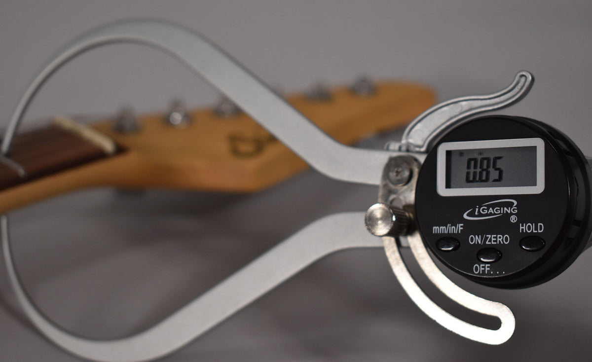 A digital caliper measures the guitar's neck, showing "0.85" on its screen, with the headstock blurred in the background.