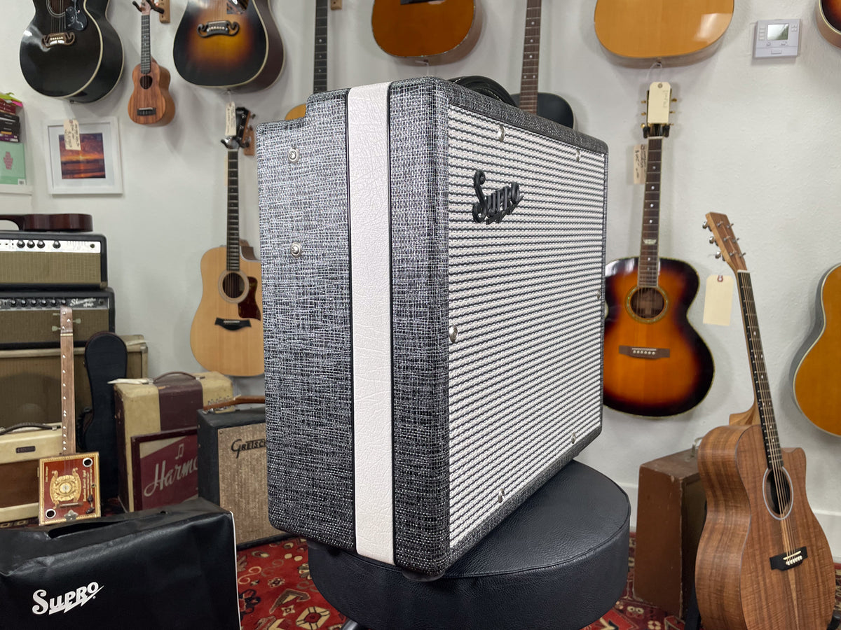A Supro 1600 Supreme combo amp with a black and white tweed finish and a black Supro logo, sitting on a black stool.
