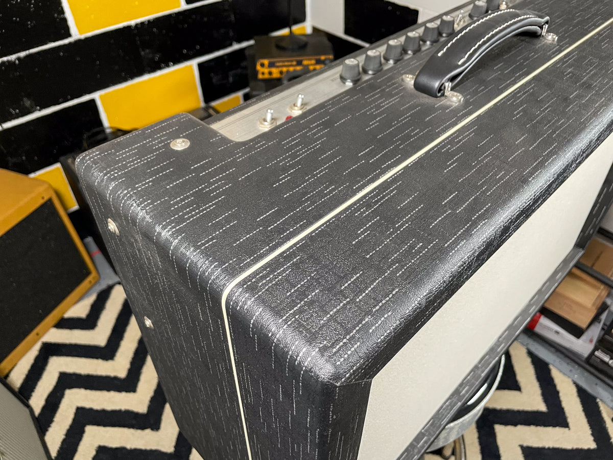 Top-down view of a black textured amp with white piping, a black leather handle, and silver control knobs on the top panel.