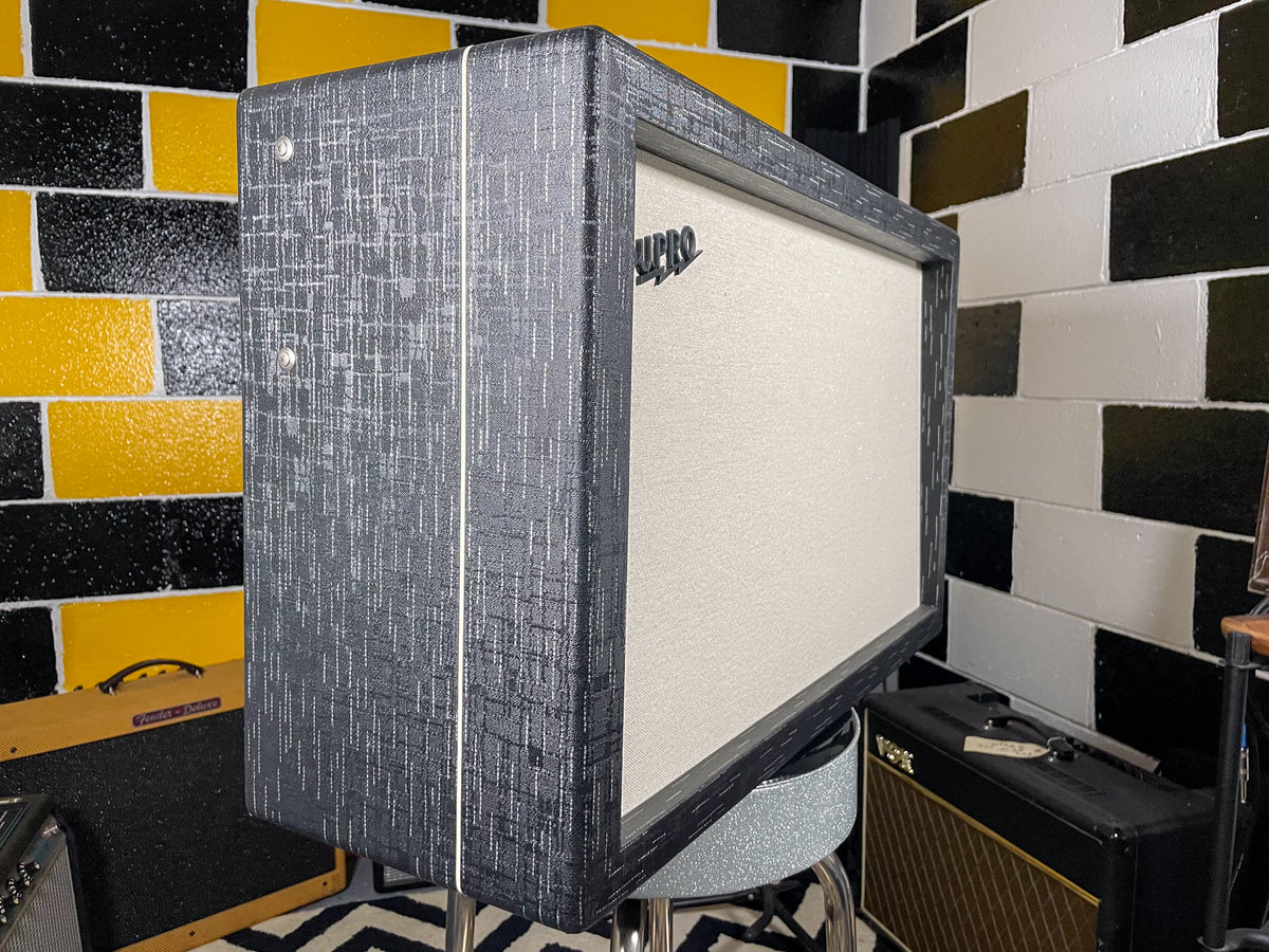 Angled view of the amp, showing its dark gray textured tolex with white piping and a light cream grille cloth with a black logo.
