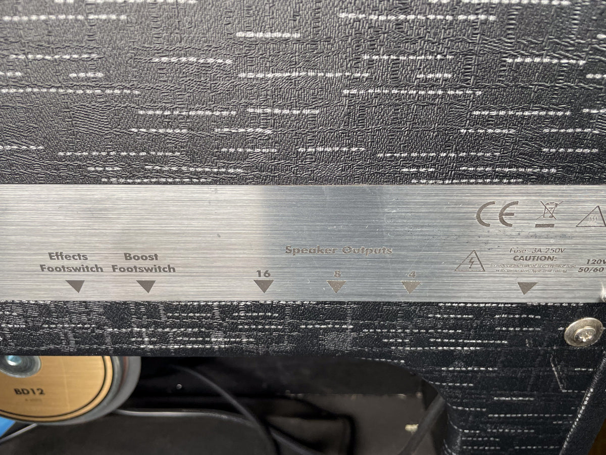 Close-up of the amp's brushed metal rear panel, showing footswitch and speaker outputs, with black textured tolex above and below.